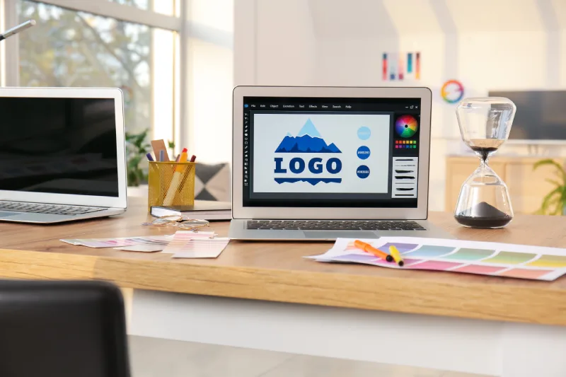 A designer's workspace featuring a laptop displaying a mountain-themed logo design, color swatches, and an hourglass on a wooden desk.
